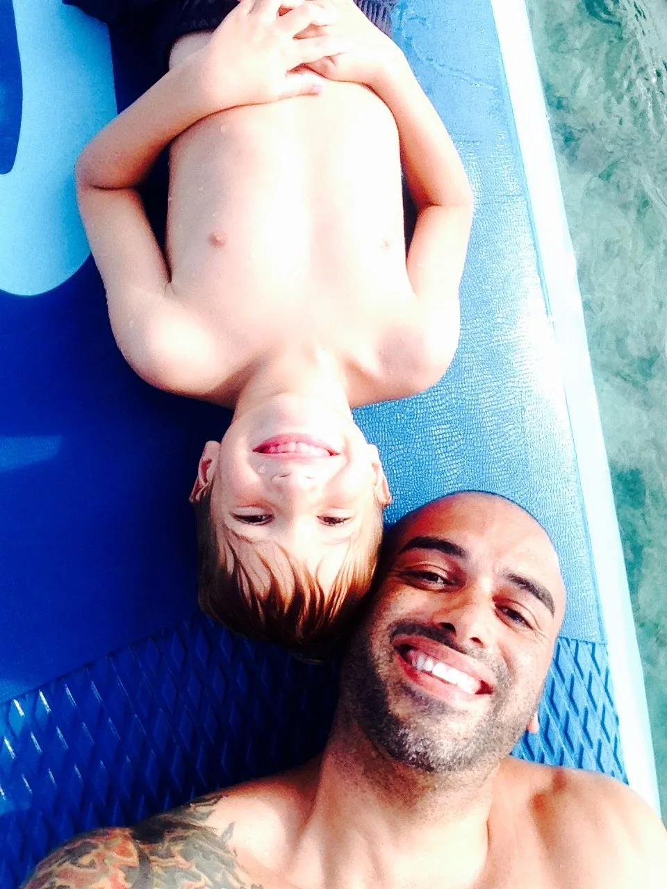Father and son lying head to head on a blue paddleboard, smiling at the camera
