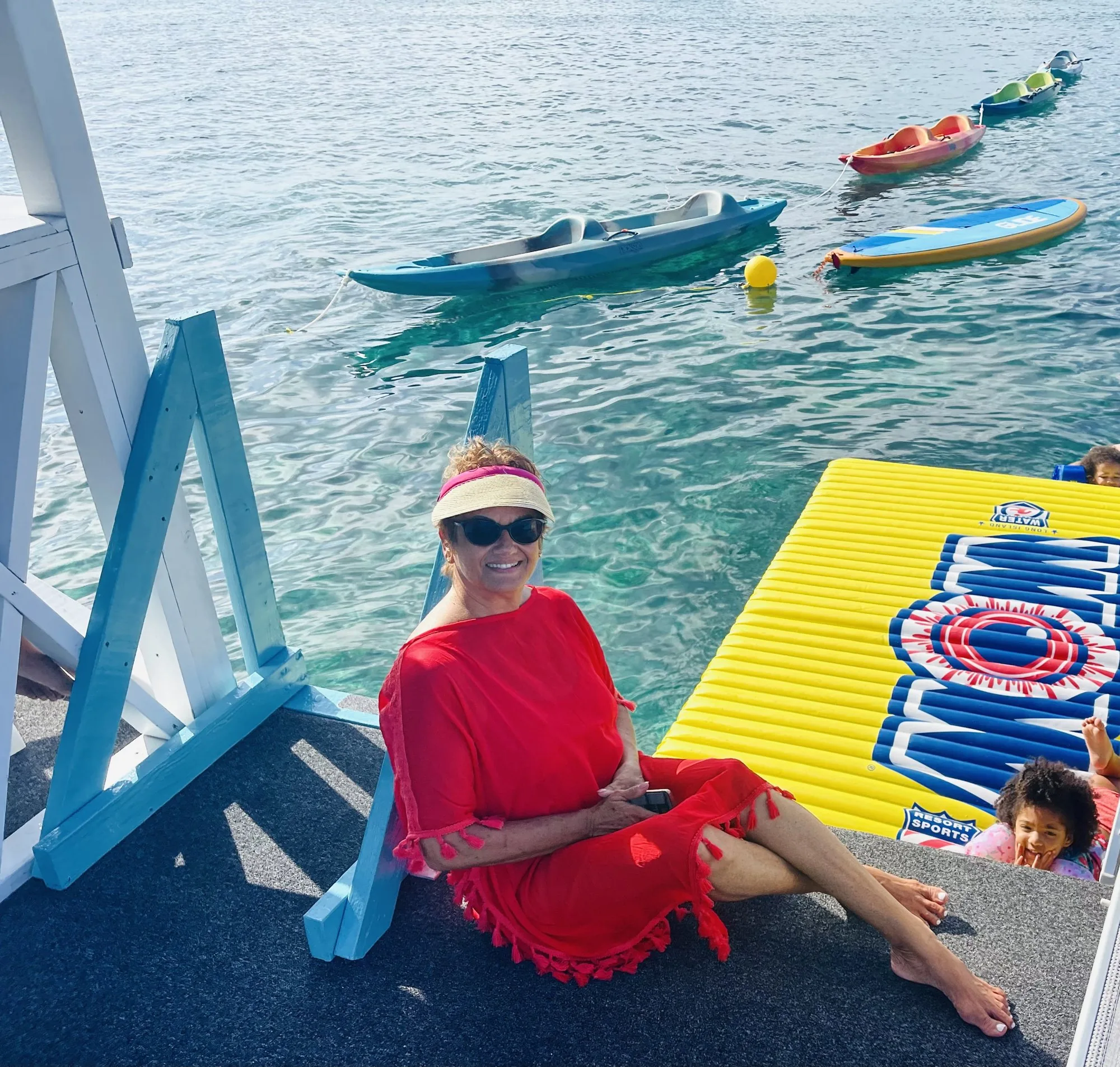 Woman in a red dress sitting on a dock above kayaks, a paddleboard and a yellow water mat
