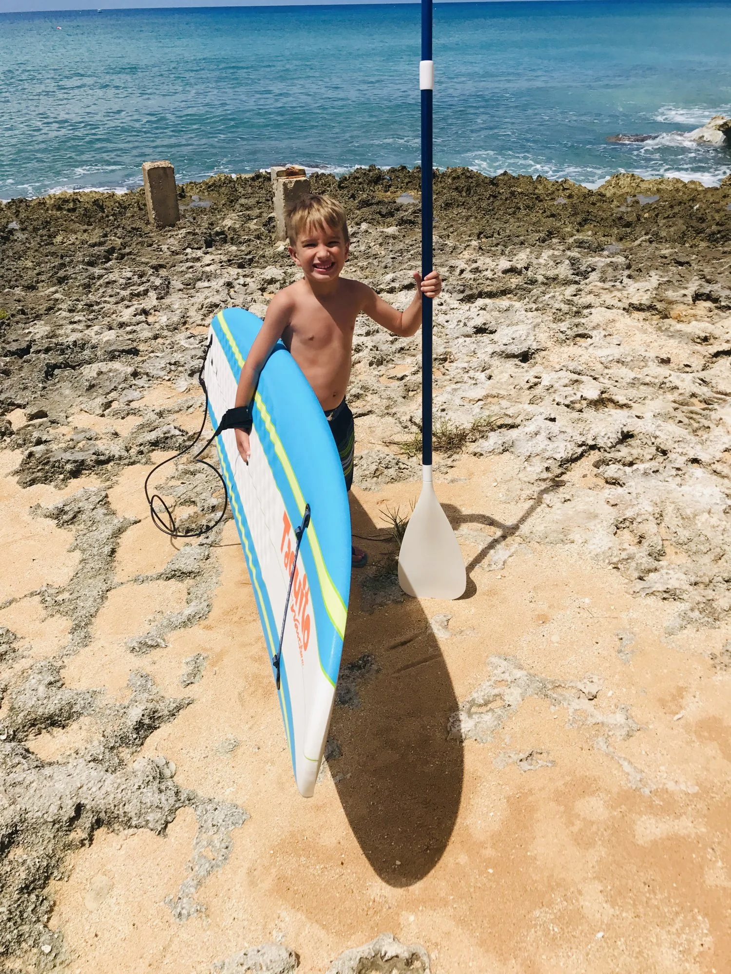 Young boy carrying a paddleboard and paddle on the ironshore by the sea