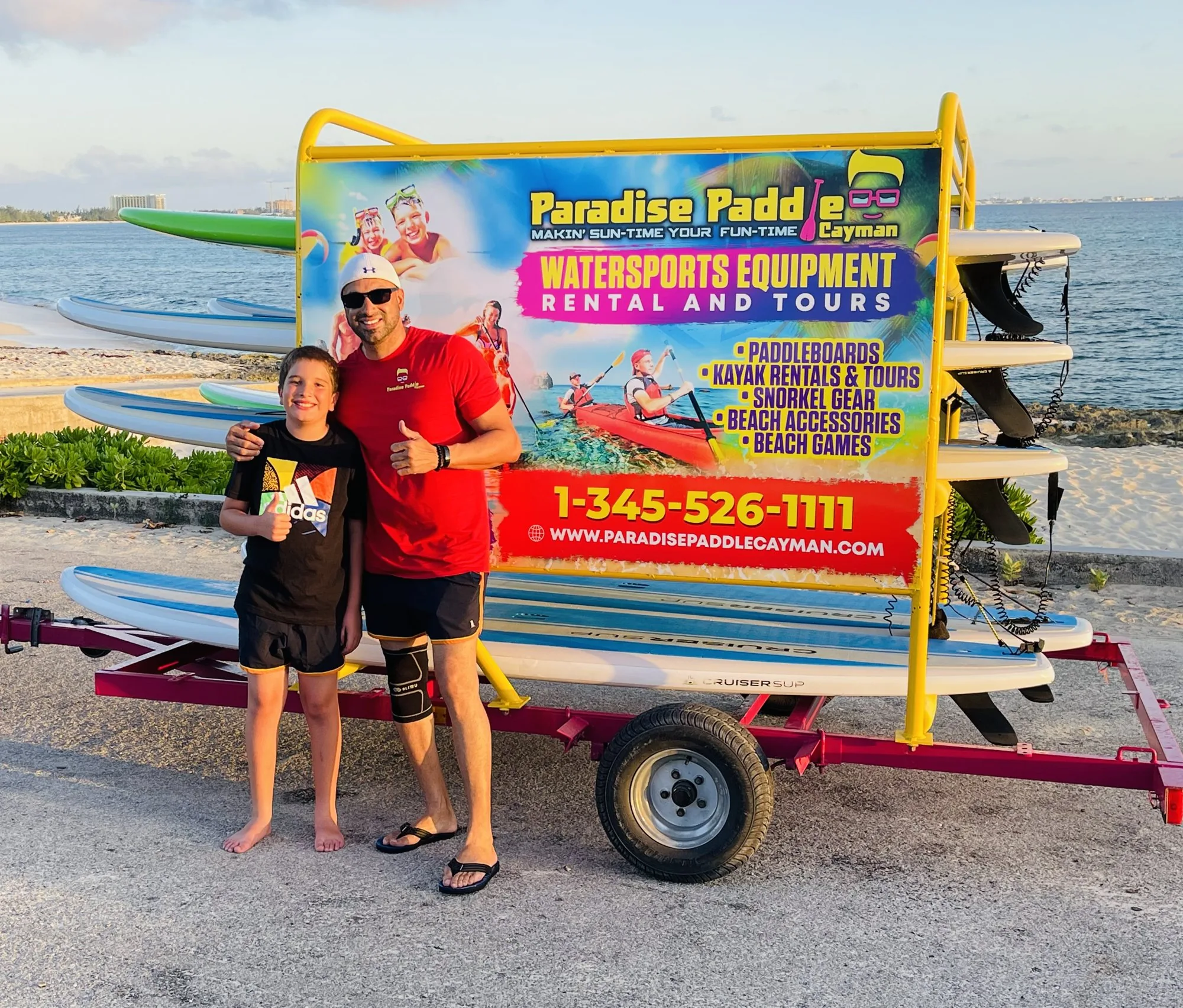 Father and son giving a thumbs up in front of the Paradise Paddle paddleboard trailer by the sea