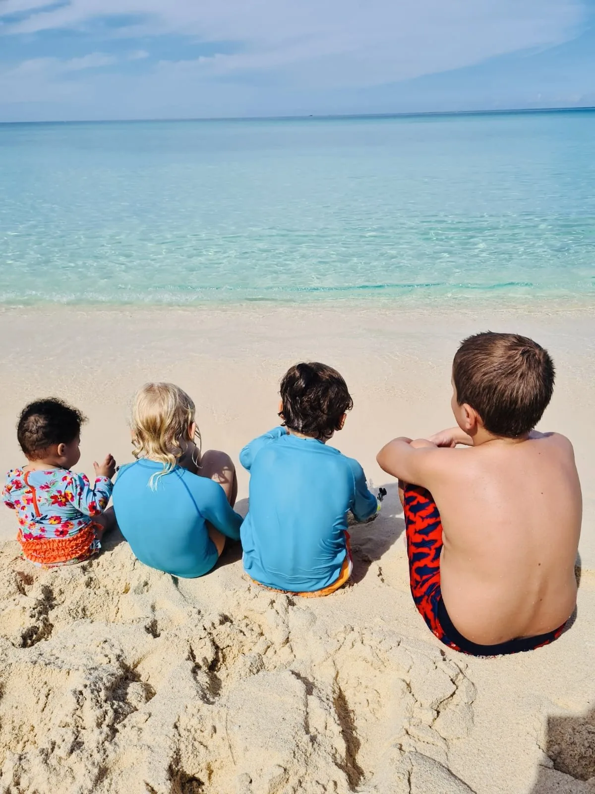 Four children sitting on the sand looking out at the turquoise sea