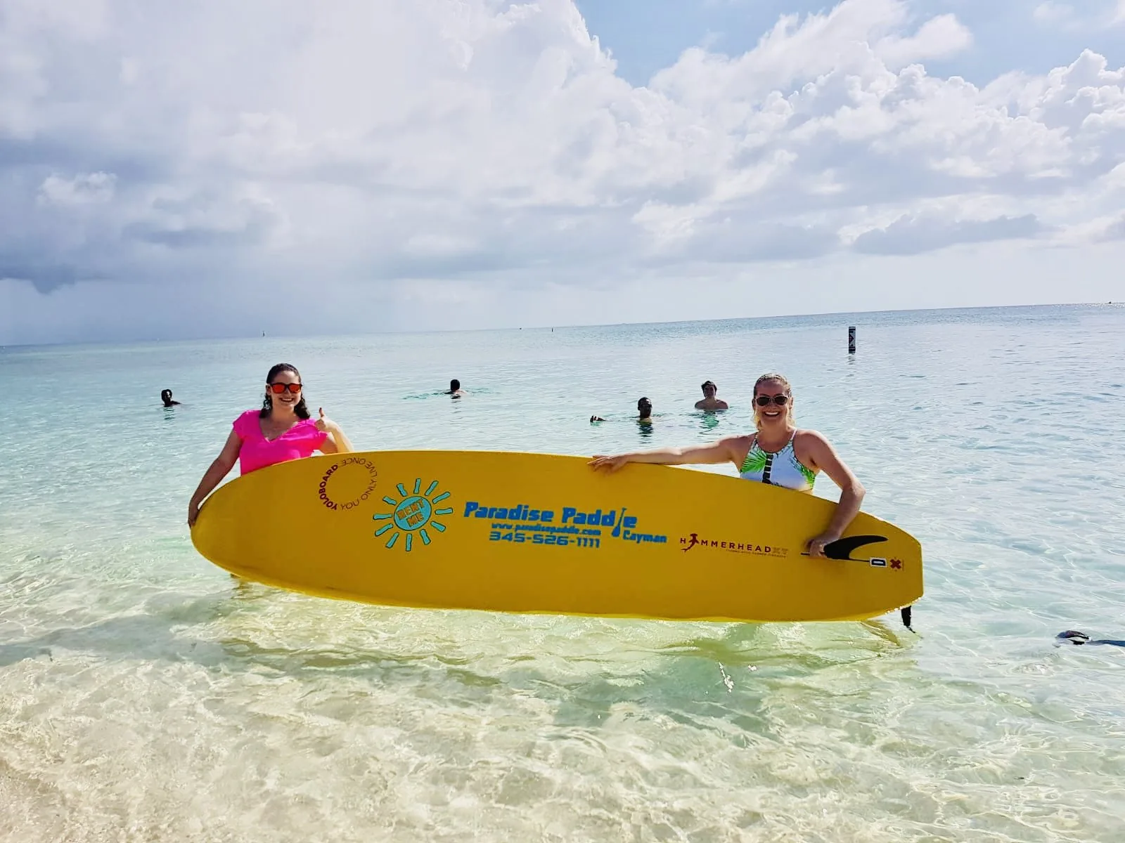 Two women holding a yellow Paradise Paddle board in the shallow turquoise water