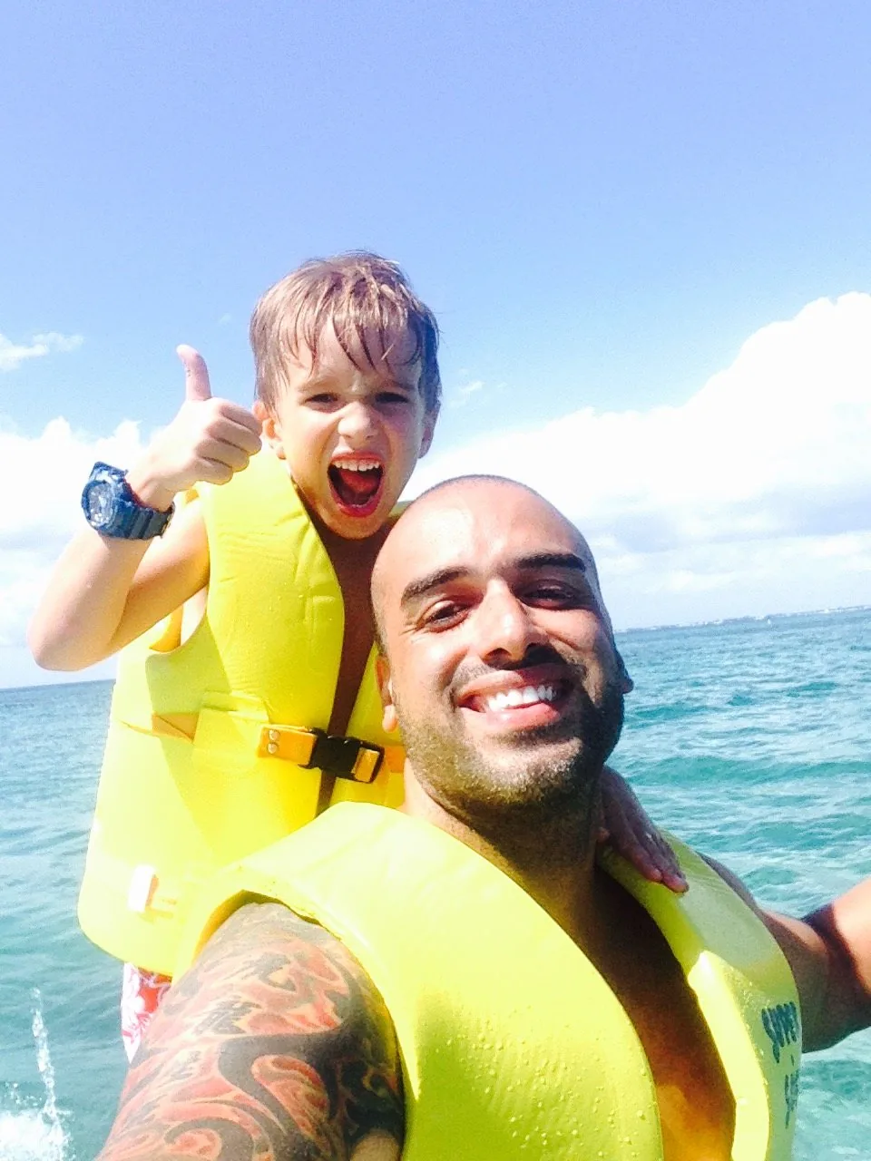 Boy in a yellow life jacket giving a thumbs up beside his father out on the water