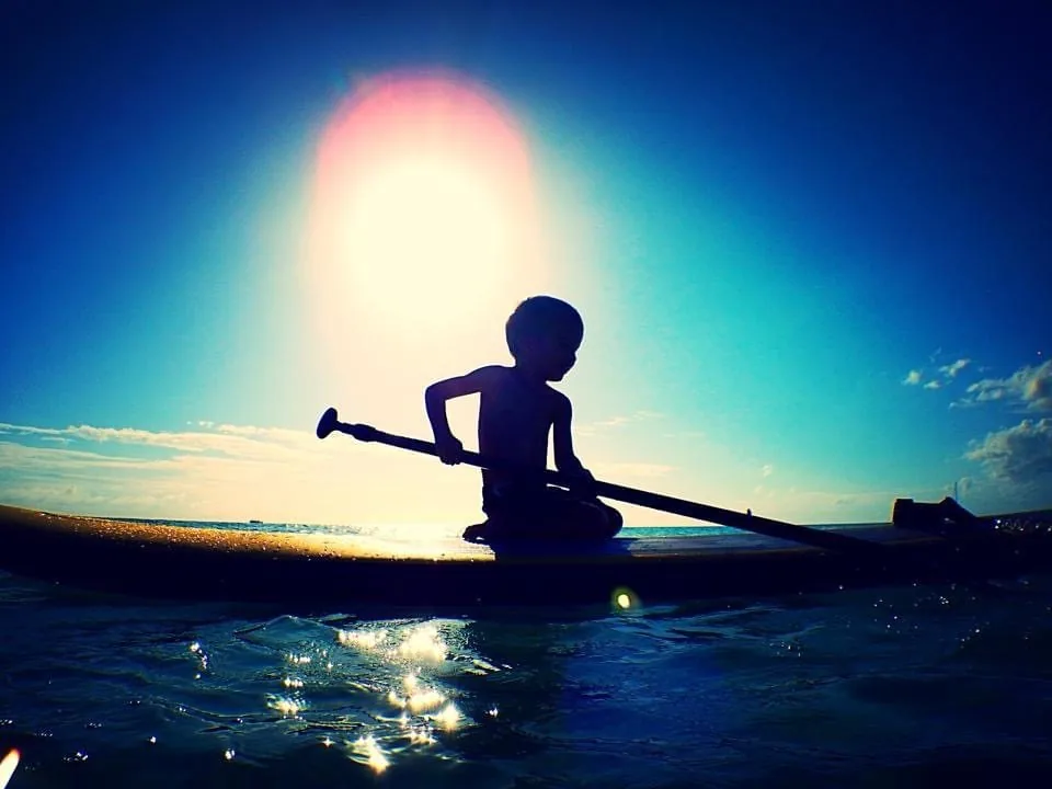 Silhouette of a young boy paddling a board under a bright sun